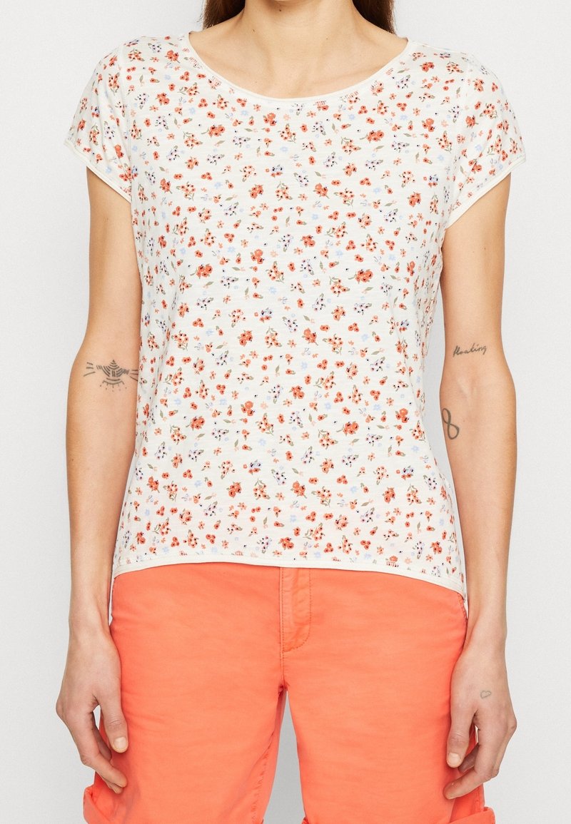 Femme portant un haut blanc à manches courtes à motif floral avec un short orange, montrant des tatouages sur les deux avant-bras et les mains.