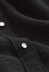 Chemise noire en tissu avec une surface texturée ; possède des boutons ronds en métal doré le long de l'avant, mettant en valeur un design lisse et minimal.