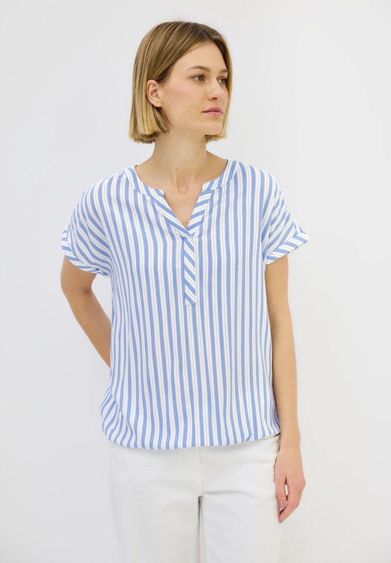 Blusa a maniche corte blu e bianca a righe con scollo a V e design morbido, realizzata in tessuto leggero. Abbinata a pantaloni bianchi.
