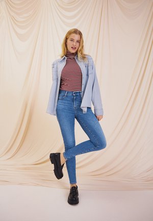 Veste en denim clair sur un col roulé rayé rouge et blanc, associée à un jean skinny bleu et des chaussures noires chunky sur un fond neutre.