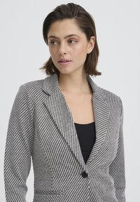 Blazer a righe in bianco e nero; presenta un rever a lancia, chiusura con un solo bottone e tasche frontali con tessuto strutturato.