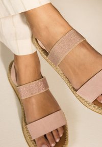 Sandales en daim rose avec deux larges bandes, dont une avec un accent pailleté. Semelle en jute ajoutant de la texture. Vue en gros plan, pied élégamment positionné.