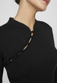 Črna pletena majica z mandarinskim ovratnikom, asimetričnim dizajnom in dekorativnimi gumbi ob stranski odprtini.