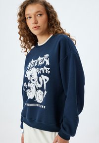 Sudadera azul marino de tejido suave, con un estampado gráfico blanco de un osito de peluche y texto. Manga larga con puños acanalados.
