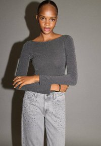 Siva dolgi rokav v kovinskem pletenju z širokim čolnskim izrezom, v kombinaciji s svetle sivimi džinsih, okrašenimi z diamanti na žepih.