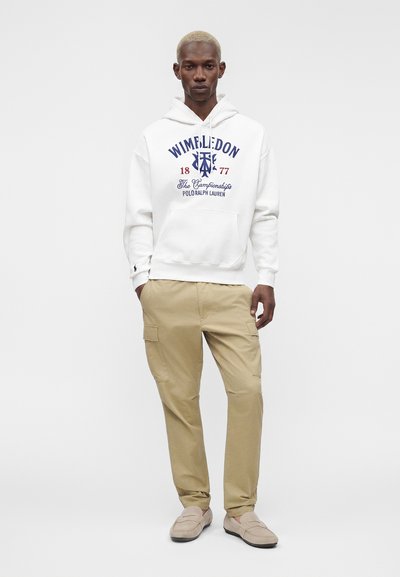 Άνδρας με λευκή μπλούζα με κουκούλα Wimbledon Polo Ralph Lauren, μπεζ cargo παντελόνια και μπεζ μοκασίνια, που στέκεται μπροστά σε ένα ομοιόμορφο ανοιχτό γκρι φόντο.