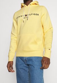 Sudadera amarilla hecha de una mezcla de algodón, con un bolsillo frontal, capucha con cordón y texto del logo en negro con el año de establecimiento. Logo adicional en la manga.