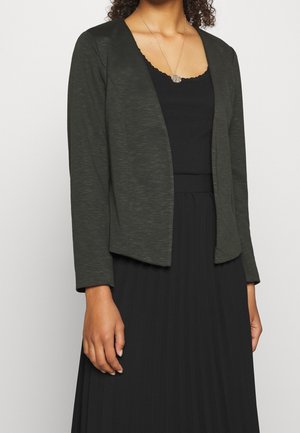 Nainen, joka pukee tummanvihreän avoimen jakkupuvun, mustan pitsireunuksella koristellun ribbineuletopin, mustan laskostetun hameen ja pyöreän riipuskaulakorun.