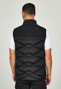 Črn puffer brezrokavnik z diamanti vzorcem, visokim ovratnikom in gladko teksturo. Nosite ga čez belo majico, kar prikazuje sproščen fit.