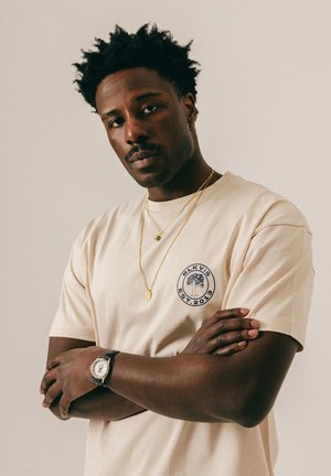 Man met gekruiste armen, een beige t-shirt met een circulair boomlogo, gouden kettingen, een creooloorbel en een polshorloge, die recht vooruit kijkt.