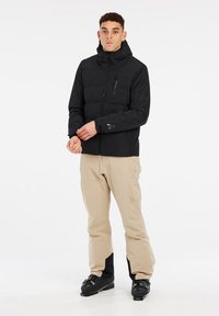 Chaqueta aislante negra con capucha, bolsillo en el pecho con cremallera, combinada con pantalones beige y botas negras, sobre un fondo blanco.