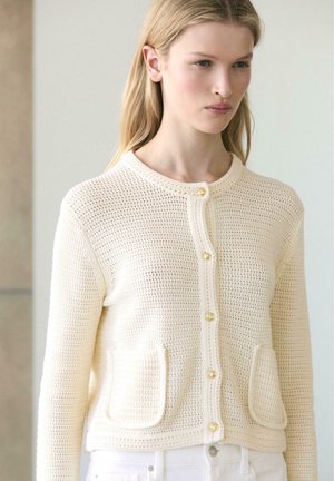 Giovane donna che indossa un cardigan lavorato a maglia color crema con bottoni dorati e tasche anteriori, abbinato a pantaloni bianchi, sfondo neutro.