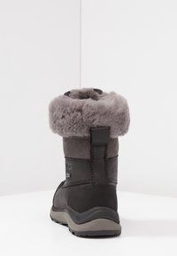 Czarny zimowy but z szarym futrem z owczej skóry, teksturowana skóra i gumowa podeszwa, widoczny z tyłu na białej powierzchni.