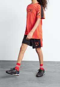 Niño caminando vestido con ropa deportiva CR7 de Nike en rojo y negro, con calcetines rojos y botas de fútbol negras, sobre un suelo gris contra una pared blanca.