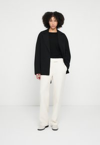 Blazer negru supradimensionat cu reveruri decupate, purtat peste un top negru mulat și pantaloni largi cu textură crem, asortați cu pantofi deschisi la culoare.