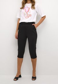 Biała koszulka graficzna z kolorową ilustracją napoju, zestawiona z czarnymi spodniami w kant o 7/8 długości oraz bocznymi kieszeniami. Czarny obcas sandały.