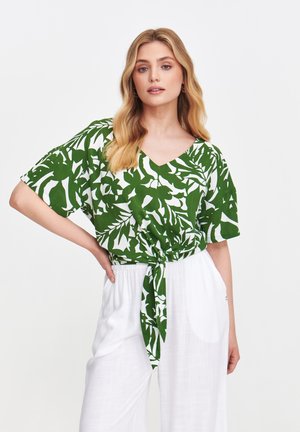 Zielona i biała bluzka w wzory z dekoltem w kształcie litery V i krótkimi rękawami, z przodu z węzłem. W połączeniu z białymi spodniami o szerokich nogawkach.