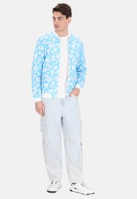 Jachetă bleu deschis cu fermoar, cu modele florale albe, asociată cu un tricou alb și pantaloni cargo bleu deschis. Încălțămintea are accente negre.