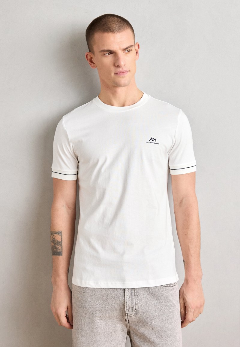 T-shirt bianca con collo rotondo e maniche corte, caratterizzata da un bordo nero sulle maniche e un piccolo logo sul petto. Materiale in cotone morbido.