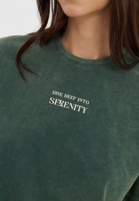 Tamsiai žalia džemperis su minkštu, tekstūruotu paviršiumi ir baltu tekstu, kuris sako "NARDYK DEGLYJE" per krūtinę.