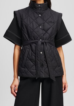 Kvinde iført en sort quiltet ærmeløs vest med trykknapper og taljebånd over en sort kortærmet top og sorte bukser.