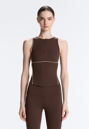 Štíhla žena oblečená v hnedom bezrukávovom topán so zipsom vpredu a zodpovedajúcich priliehavých nohaviciach, stojaca na jednoduchom svetlom pozadí.