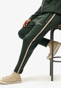 Grønne sweatpants med beige og marineblå sidestriber, prydet med "Superdry"-logoet. Båret med beige højtop sneakers og siddende på en metalstol.