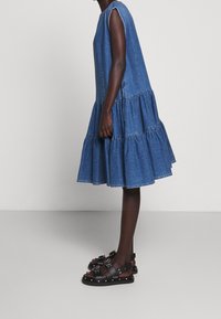 Denim šatové šaty bez rukávů s vrstveným volánem na spodním lemu, v středně modré barvě. V kombinaci s černými sandály s hroty, které mají více popruhů a spon.