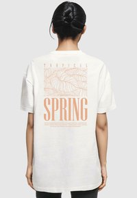 Cremefarvet oversized t-shirt med et grafisk design af tropiske blade og ordet "SPRING" i store lys orange bogstaver på ryggen.