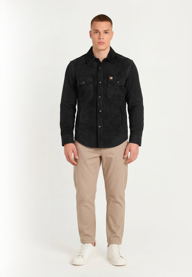 Young man standing, wearing a black buttoned jacket with chest pockets, beige trousers, and white sneakers, against a plain light background.