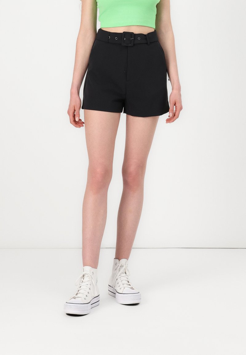 Svarte skreddersydde shorts med en tettsittende midje og beltesløyfer, med et matchende belte. Kombinert med hvite høye sneakers og en grønn topp.