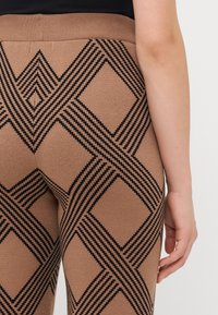 Bruine gebreide legging met een zwart diamantpatroon, een gestructureerde afwerking en een brede tailleband. Zichtbare hand die op de stof rust.