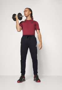 Burgundy tričko s krátkym rukávom a čiernym logom Adidas, v kombinácii s čiernymi teplákovými nohavicami a čiernymi teniskami s červenými akcentmi, držiac jednoručný závažie.