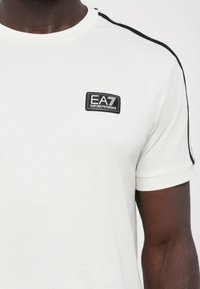 White cotton t-shirt with black side stripes, ribbed crew neck, and a rectangular EA7 Emporio Armani logo on the chest. Short sleeves.