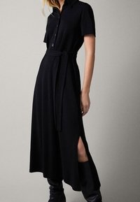 Sort, knælængde kjole med krave, korte ærmer, knapper, sideslids og et matchende bælte til justering af pasformen.