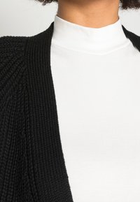 Černý pletený kardigan s texturovaným vzorem, s otevřeným předním dílem, nošený přes bílý top s vysokým límcem.