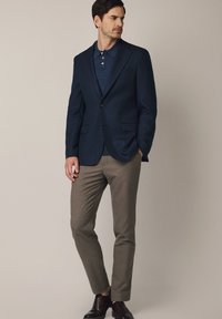 Granatowa marynarka z pojedynczym guzikiem, dwoma kieszeniami z przodu, zestawiona z ciemnoniebieskim polo i jasnobrązowymi spodniami typu slim-fit. Brązowe skórzane buty dopełniają całość.