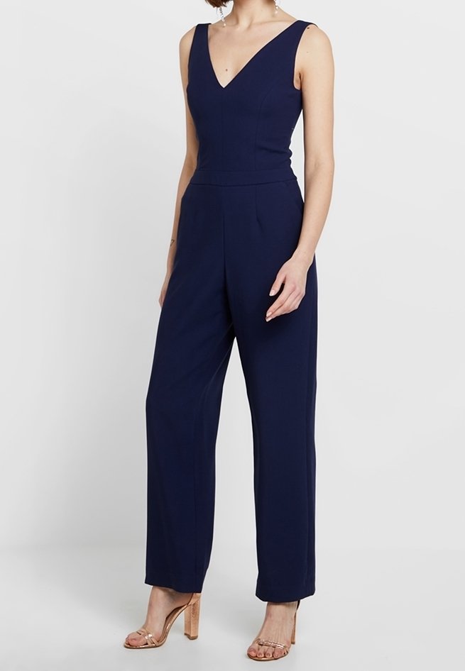 Femme portant une combinaison bleu marine sans manches avec un pantalon à jambes larges et un décolleté en V, associée à des sandales à talons hauts couleur nude.