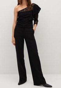 Top nero a spalla unica con dettagli in pizzo e ruches; abbinato a pantaloni neri a vita alta con gamba larga e scarpe con punta.