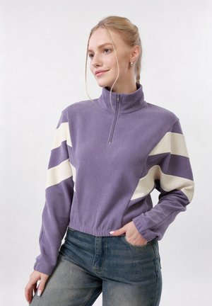 Νεαρή γυναίκα με ξανθά μαλλιά σε πλεξούδα φοράει μοβ και λευκό πουλόβερ με φερμουάρ και μπλε τζιν, ποζάροντας με το ένα χέρι στην τσέπη.