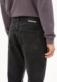 Schwarze Denim-Jeans mit einem ausgebleichten Finish, ausgestattet mit zwei hinteren Taschen und einem Etikett mit der Aufschrift "ARMEDANGELS" am Bund. Strukturiertem Stoff.