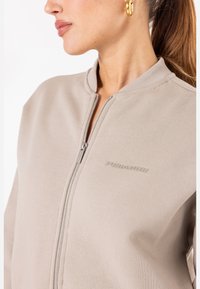 Lys beige zip-up jakke med ribbestrikket krage, med preget logo på brystet og glatt tekstur. Glidelås foran for lukking.