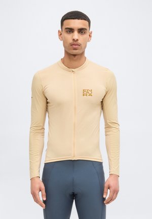 Ung mann iført en beige sykkeltrøye med lang arm og frontglidelås, samt marineblå polstrede sykkelshorts, stående mot en ensfarget lys bakgrunn.