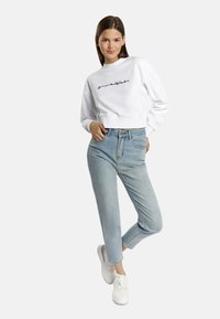 Biała, krótka bluza z haftowanym napisem, zestawiona z jasnoniebieskimi dżinsami. Białe sneakersy dopełniają stylizację, posiadając gładką teksturę.