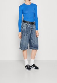 Maglia a maniche lunghe blu a coste, pantaloni shorts in denim larghi con tasche, cintura nera, calzini bianchi e sneakers nere con strisce bianche.