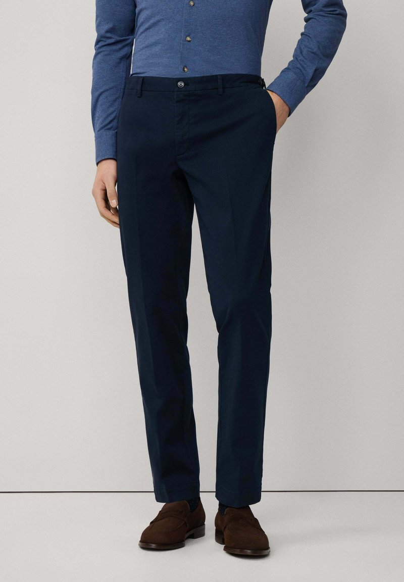 Dunkelblaue Hose aus glattem Stoff mit flachem Bund, Gürtelschlaufen und geraden Beinen, kombiniert mit braunen Slippern.