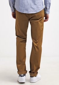 Braune Baumwollchinos mit geradem Schnitt, umgeschlagenen Säumen und glatter Textur, kombiniert mit weißen Turnschuhen. Hellblaues Hemd sichtbar.