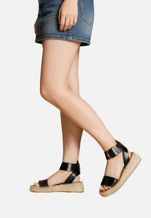 Jambes portant des sandales compensées espadrilles en cuir noir avec bride à la cheville et une courte jupe en denim bleu sur fond blanc.