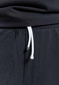 Shorts de sport en mesh noir avec cordon blanc et une ceinture élastique noire sous l’ourlet d’un t-shirt noir.