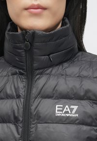 Melnā pūkaina jaka ar augstu apkakli, horizontālu šuvi, priekšējo rāvējslēdzēju un baltu EA7 logo uz krūtīm. Gluda, viegla auduma.